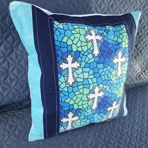 May include: Decorative pillow with a stained glass design featuring white crosses on a blue and green mosaic background. The pillow has a navy blue border and a light blue side panel.