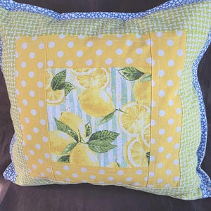 May include: Square decorative pillow with a lemon theme. The pillow features a central panel with lemon slices and leaves on a striped background. It has a yellow polka dot border and a green and blue patterned edge.