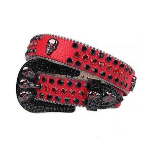 Puede incluir: Cinturón de cuero rojo con hebilla y adornos de calavera negros. El cinturón está tachonado con pedrería negra y roja. La hebilla y las trabillas son negras.