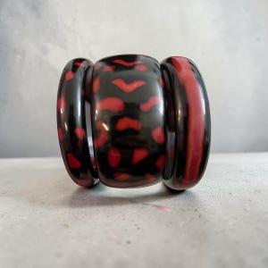 May include: A wide, chunky bracelet with a bold design. The bracelet features a glossy black base with red, irregular shapes and stripes. The bracelet is made of a hard, smooth material and is set against a neutral background.