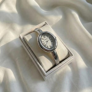 Puede incluir: Un reloj de pulsera plateado con esfera ovalada y n&uacute;meros romanos. El reloj, de la marca "NAIDU", se presenta en una caja blanca, sobre un fondo de tela blanca y suave. La correa del reloj tiene un dise&ntilde;o elegante y minimalista.