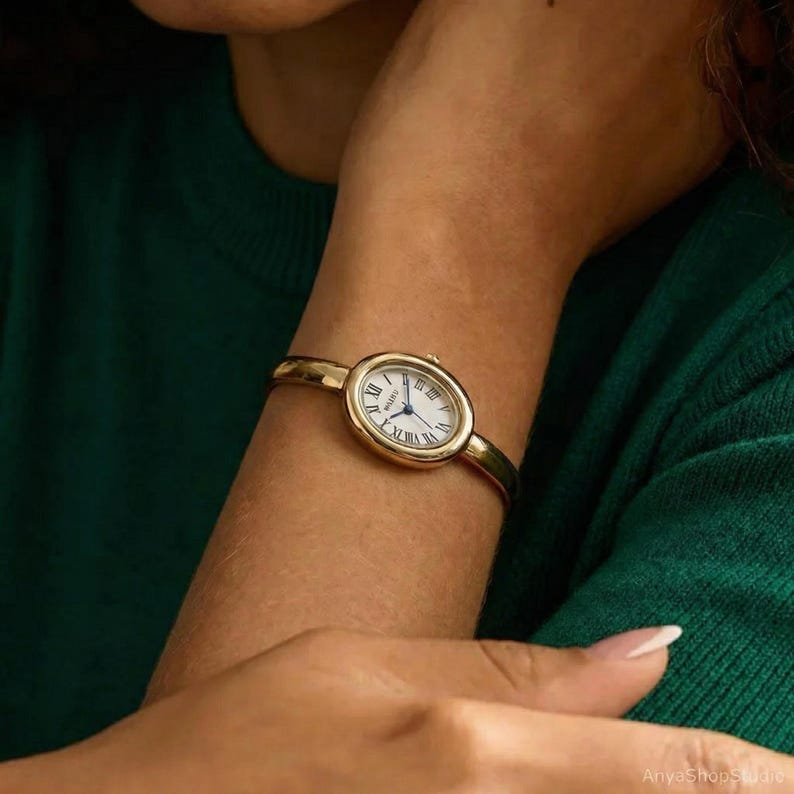 Puede incluir: Un reloj de pulsera dorado con esfera blanca y n&uacute;meros romanos. El reloj tiene una banda delgada y una esfera ovalada. El reloj se lleva en la mu&ntilde;eca, y el fondo es verde oscuro.