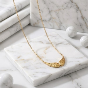 May include: A gold-colored necklace featuring a pair of stylized wings. The wings are detailed with etched lines, and the necklace chain is delicate. The jewelry rests on a white marble surface, with two white marble spheres in the background.