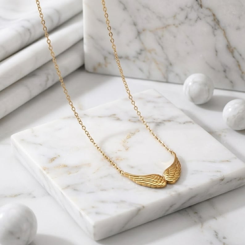 May include: A gold-colored necklace featuring a pair of stylized wings. The wings are detailed with etched lines, and the necklace chain is delicate. The jewelry rests on a white marble surface, with two white marble spheres in the background.