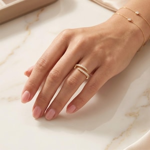 Op de afbeelding: Een goudkleurige ring met een dubbelbandontwerp, bezet met kleine, sprankelende stenen. De ring wordt gedragen op een hand, met een bijpassende gouden armband met kleine, ronde details. De achtergrond is licht en neutraal.