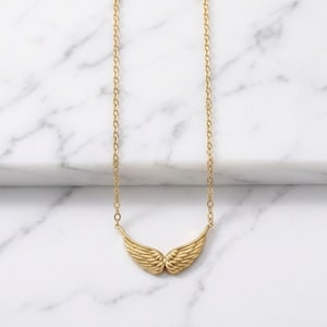 May include: A gold-colored necklace featuring a pair of detailed wings. The wings are the focal point, suspended from a delicate chain. The necklace is displayed against a white marble background, highlighting the jewelry's design.
