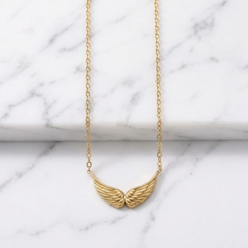 May include: A gold-colored necklace featuring a pair of detailed wings. The wings are the focal point, suspended from a delicate chain. The necklace is displayed against a white marble background, highlighting the jewelry's design.