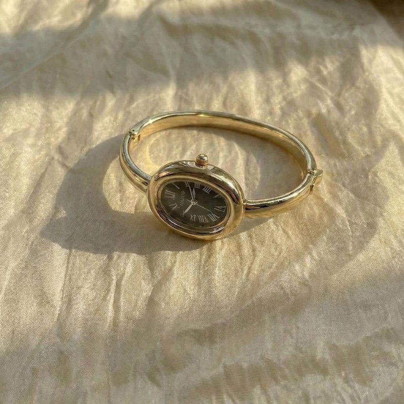 Puede incluir: Reloj de pulsera dorado con esfera ovalada y n&uacute;meros romanos. El reloj tiene una esfera oscura y un dise&ntilde;o de pulsera con bisagras. El reloj se muestra sobre una superficie texturizada de color crema.