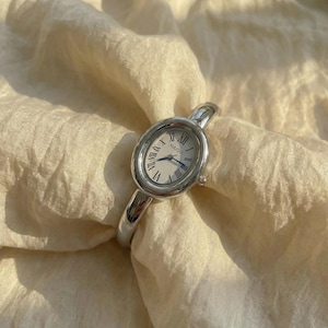 Puede incluir: Un reloj de pulsera plateado con esfera ovalada y n&uacute;meros romanos. La correa del reloj es de estilo brazalete plateado a juego. La esfera del reloj tiene la marca "NAIDU" impresa. El reloj se muestra sobre una tela color crema.