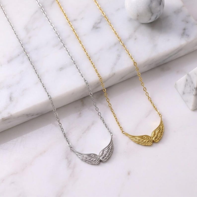 May include: Two necklaces with wing pendants. One necklace is silver, the other is gold. The pendants are detailed with feather-like textures. The chains are delicate and the background is a white marble surface.
