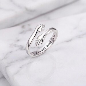 Könnte beinhalten: Ein silberner Ring mit zwei Armen, die sich umarmen. Der Ring trägt die Inschrift "Always with you". Der Ring liegt auf einer weißen Marmoroberfläche.