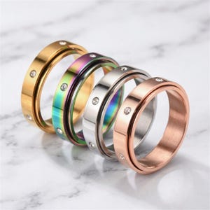 Peut inclure: Quatre bagues rotatives en or, irisé, argent et or rose. Chaque bague a une bande avec un centre rotatif et de petits pierres précieuses transparentes. Les bagues sont présentées sur une surface en marbre blanc.