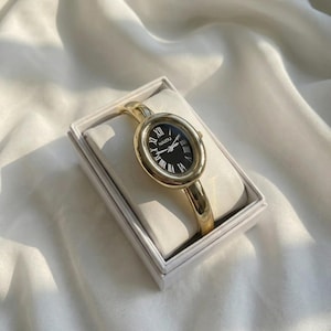 Puede incluir: Reloj de pulsera ovalado dorado con esfera negra y n&uacute;meros romanos. El reloj se muestra en una caja blanca sobre una superficie de sat&eacute;n blanco. La correa del reloj es de estilo brazalete dorado.