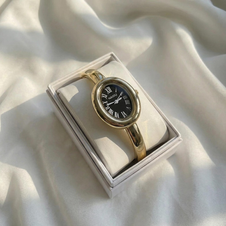 Puede incluir: Reloj de pulsera ovalado dorado con esfera negra y n&uacute;meros romanos. El reloj se muestra en una caja blanca sobre una superficie de sat&eacute;n blanco. La correa del reloj es de estilo brazalete dorado.
