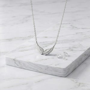 May include: A silver-toned necklace with a wing pendant and a delicate chain. The pendant features detailed feathering. Two small, clear gemstones are also present, all displayed on a white marble surface. Jewelry.