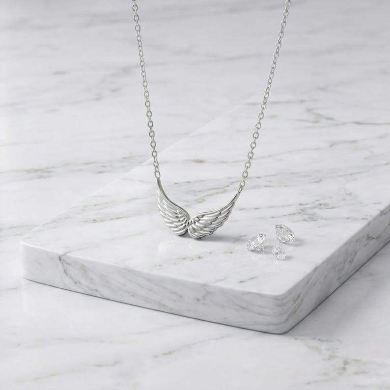 May include: A silver-toned necklace with a wing pendant and a delicate chain. The pendant features detailed feathering. Two small, clear gemstones are also present, all displayed on a white marble surface. Jewelry.