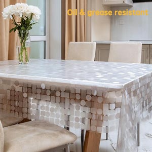 May include: A clear, rectangular plastic tablecloth featuring a repeating pattern of white circles and squares. The text "Oil & grease resistant" is printed in yellow at the top. The tablecloth covers a dining table, with a glass vase of white flowers.