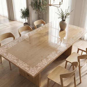 May include: A rectangular wooden dining table featuring a clear protective cover with a white intricate floral lace-style border pattern. The table is surrounded by six light-toned wooden chairs, and a ceramic vase with greenery sits on top.