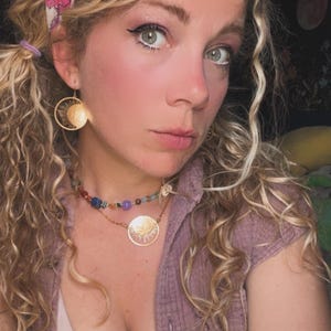 Puede incluir: Una mujer con cabello rubio rizado que lleva una diadema floral rosa, aretes dorados con forma de sol y un collar de piedras preciosas multicolores. Tambi&eacute;n lleva una camiseta rosa y una camisa abotonada de color p&uacute;rpura claro.