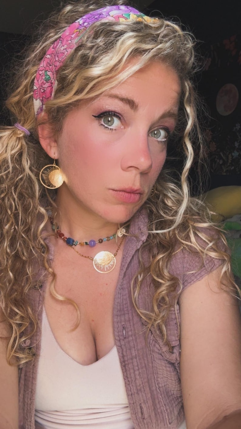 Puede incluir: Una mujer con cabello rubio rizado que lleva una diadema floral rosa, aretes dorados con forma de sol y un collar de piedras preciosas multicolores. Tambi&eacute;n lleva una camiseta rosa y una camisa abotonada de color p&uacute;rpura claro.
