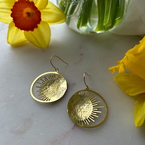 Puede incluir: Pendientes dorados con dise&ntilde;o de sol. Cada pendiente presenta un disco dorado martillado con un patr&oacute;n similar al sol recortado en el borde. Los pendientes se exhiben sobre una superficie de m&aacute;rmol, con narcisos amarillos en el fondo.