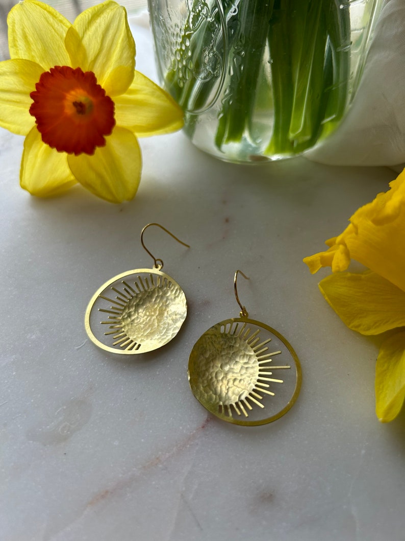 Puede incluir: Pendientes dorados con dise&ntilde;o de sol. Cada pendiente presenta un disco dorado martillado con un patr&oacute;n similar al sol recortado en el borde. Los pendientes se exhiben sobre una superficie de m&aacute;rmol, con narcisos amarillos en el fondo.