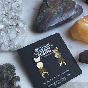 May include: A pair of gold-toned drop earrings featuring a vertical arrangement of three hammered circular and crescent moon shapes. The earrings are displayed on a black card printed with the text EMBODY AURA, LET YOURSELF SHINE, HYPOALLERGENIC STAINLESS STEEL, WWW.EMBODYAURA.ETSY.COM, and @EMBODY_AURA.