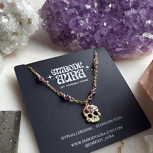 Puede incluir: Collar dorado con un colgante de calavera adornado con piedras preciosas rojas y moradas. El collar se exhibe en una tarjeta negra con el texto "EMBODY AURA" y "LET YOURSELF SHINE". Cristales de amatista en el fondo.