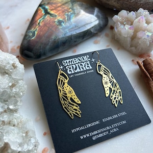 May include: Gold-tone earrings featuring a hand design and the text "embody aura let yourself shine". The earrings are hypoallergenic and made of stainless steel. The text "www.embodyaura.etsy.com @embody_aura" is also included.