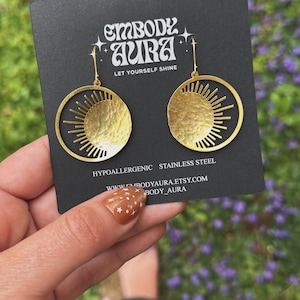 Puede incluir: Pendientes dorados con dise&ntilde;o de sol. Cada pendiente presenta un dise&ntilde;o de sol de metal martillado y texturizado dentro de un marco circular. Los pendientes se muestran en una tarjeta negra con el texto "EMBODY AURA" y "LET YOURSELF SHINE". Los pendientes son de acero inoxidable.