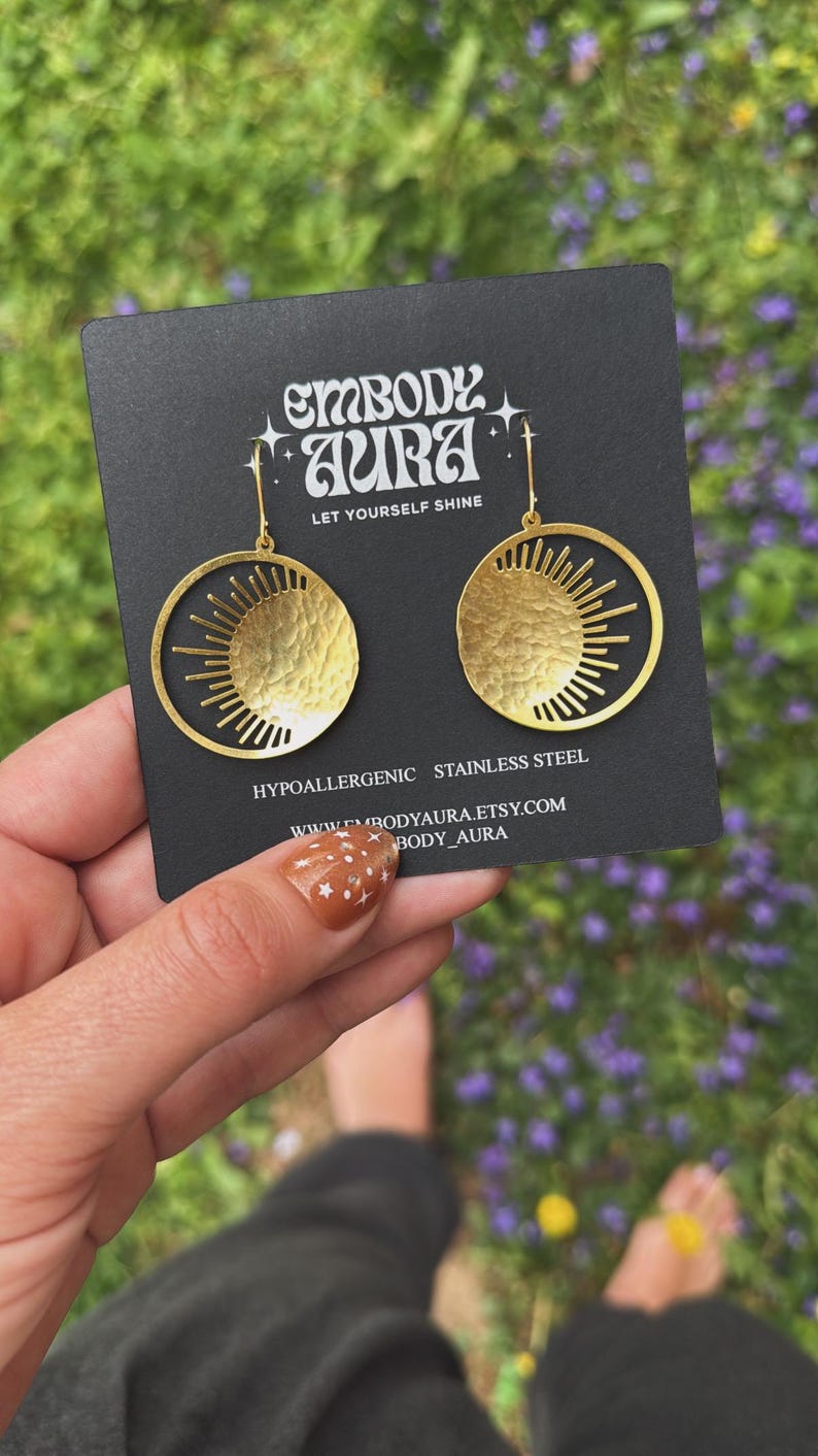Puede incluir: Pendientes dorados con dise&ntilde;o de sol. Cada pendiente presenta un dise&ntilde;o de sol de metal martillado y texturizado dentro de un marco circular. Los pendientes se muestran en una tarjeta negra con el texto "EMBODY AURA" y "LET YOURSELF SHINE". Los pendientes son de acero inoxidable.