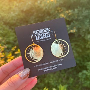 Puede incluir: Pendientes dorados con dise&ntilde;o de sol y luna. Los pendientes est&aacute;n en una tarjeta negra con el texto "EMBODY AURA" y "LET YOURSELF SHINE". La tarjeta tambi&eacute;n indica que los pendientes son hipoalerg&eacute;nicos y de acero inoxidable.