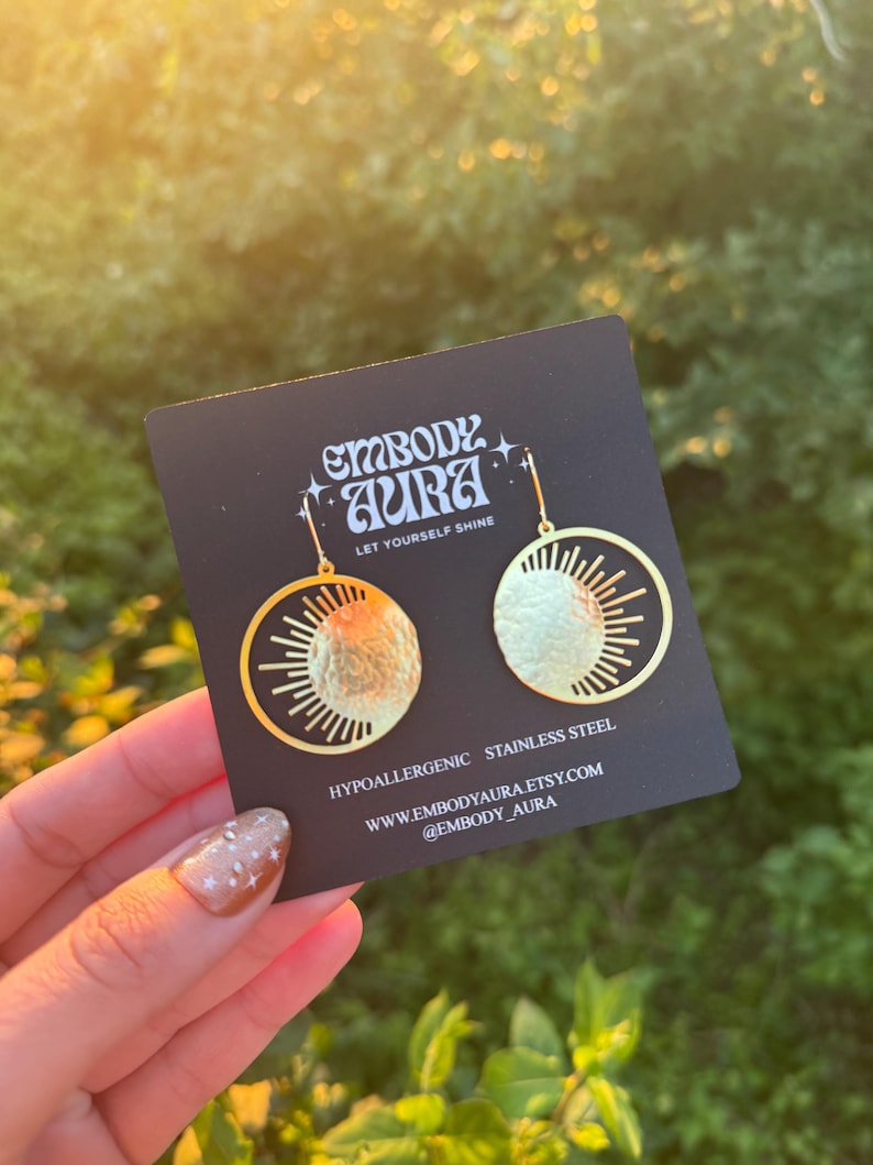 Puede incluir: Pendientes dorados con dise&ntilde;o de sol y luna. Los pendientes est&aacute;n en una tarjeta negra con el texto "EMBODY AURA" y "LET YOURSELF SHINE". La tarjeta tambi&eacute;n indica que los pendientes son hipoalerg&eacute;nicos y de acero inoxidable.