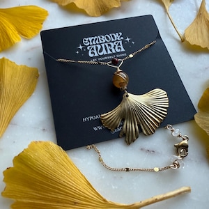May include: A gold-colored necklace with a ginkgo leaf pendant, a round amber-colored bead, and a small red bead. The necklace is displayed on a black card with the text "EMBODY AURA LET YOURSELF SHINE". Yellow ginkgo leaves surround the necklace.