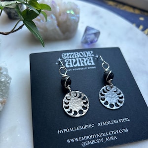 以下が含まれることがあります： 月相のデザインと黒い溶岩ビーズが付いたシルバーのイヤリングのペア。イヤリングは低刺激性で、ステンレススチール製です。カードには「Embody Aura Let Yourself Shine」と、ウェブサイトwww.embodyaura.etsy.comと@embody_auraが記載されています。