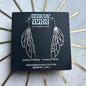 Può includere: Un paio di orecchini d'argento con un design delicato e intricato che ricorda le ali. Gli orecchini sono esposti su una carta nera con il testo "Embody Aura Let Yourself Shine" e l'indirizzo del sito web "www.embodyaura.etsy.com" e il nome del profilo sui social media "@embody_aura".