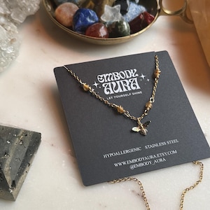 Op de afbeelding: Een goudkleurige roestvrijstalen ketting met een bijenhangertje, gepresenteerd op een zwarte kaart met de tekst "EMBODY AURA LET YOURSELF SHINE". De ketting ligt op een marmeren oppervlak, met een messing schaal met kleurrijke stenen op de achtergrond.