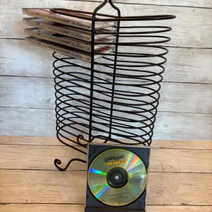 May include: A black metal wire rack designed for storing compact discs, featuring a scroll-top handle and multiple horizontal slots. Several CD cases are stacked inside the rack, and one open CD case with a LaserLight Digital disc is placed in front.