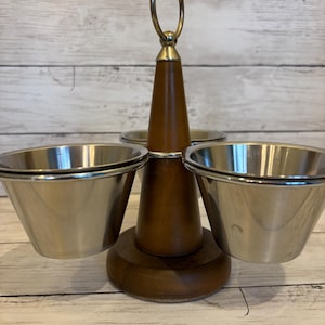 May include: A tiered serving stand featuring a central dark wood post with a circular metal handle at the top. Three removable stainless steel bowls are attached to the wooden base, arranged symmetrically around the central column for serving.