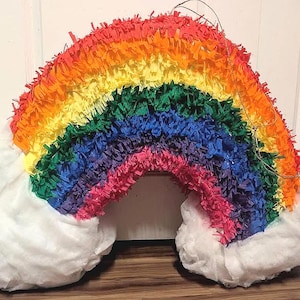 May include: A colorful rainbow piñata with layers of red, orange, yellow, green, blue, and pink fringed paper. White cotton clouds are attached to each end. The piñata is a festive decoration.