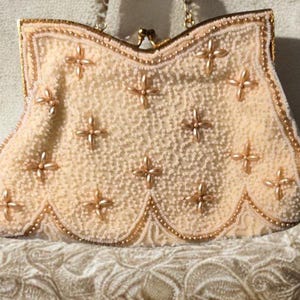 May include: Cream-colored beaded handbag with gold-tone accents. The vintage purse features a scalloped edge and is adorned with cross-shaped embellishments. The handle is a delicate chain, and the clasp is gold-toned.