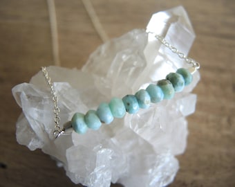 May include: A silver chain necklace with a row of light blue gemstone beads. The beads are faceted and have a slightly irregular shape.