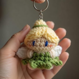 Puede incluir: Un llavero de hada de ganchillo hecho a mano. El hada tiene cabeza y pelo amarillos, ojos azules y un vestido verde. Las alas blancas están unidas a la espalda. El llavero está sujeto a un anillo de metal plateado.