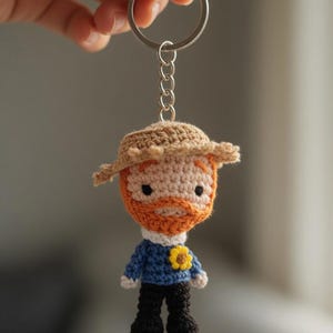 Può includere: Un portachiavi all'uncinetto fatto a mano raffigurante una figura in miniatura di Vincent van Gogh. La figura ha un viso abbronzato, barba arancione e un cappello marrone. Indossa un maglione blu con un girasole giallo e pantaloni neri. Il portachiavi è attaccato a un anello di metallo argentato.