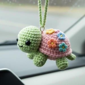 Peut inclure: Un charm de voiture en forme de tortue crochetée. La tête et les membres sont verts, la carapace rose est ornée de fleurs colorées. La tortue a des yeux noirs et une bouche souriante, suspendue par une ficelle verte.