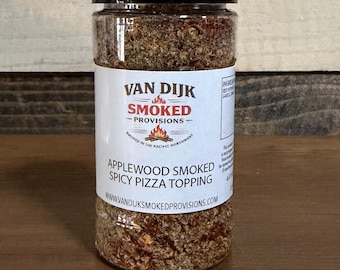 Puede incluir: Un frasco de plástico transparente con tapa negra, lleno de un aderezo para pizza granulado marrón. La etiqueta dice "VAN DIJK SMOKED PROVISIONS" y "APPLEWOOD SMOKED SPICY PIZZA TOPPING". También se ve el sitio web www.vandijksmokedprovisions.com.