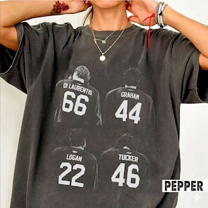 May include: A charcoal gray short-sleeve t-shirt featuring a graphic print of four individuals seen from the back, each wearing a jersey with a name and number: Di Laurentis 66, Graham 44, Logan 22, and Tucker 46. The shirt has a crew neckline.