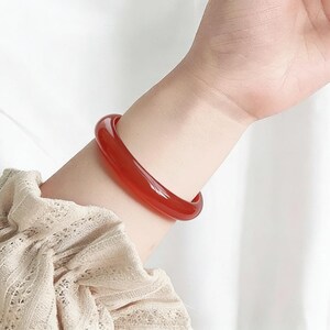 Puede incluir: Un brazalete rojo vibrante. La superficie lisa y pulida refleja la luz, mostrando el color intenso. El brazalete se lleva en la muñeca, con una manga de encaje color crema visible. Una pieza de joyería sencilla y elegante.