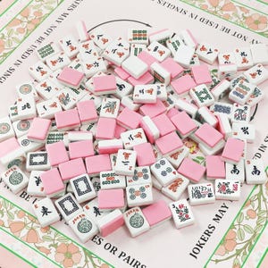 Pu&ograve; includere: Una collezione di tessere rettangolari da Mahjong con facce bianche che presentano vari simboli, caratteri e numeri tradizionali in inchiostro nero, rosso e verde, abbinate a dorsi rosa a tinta unita. Le tessere sono sparse su una superficie floreale.