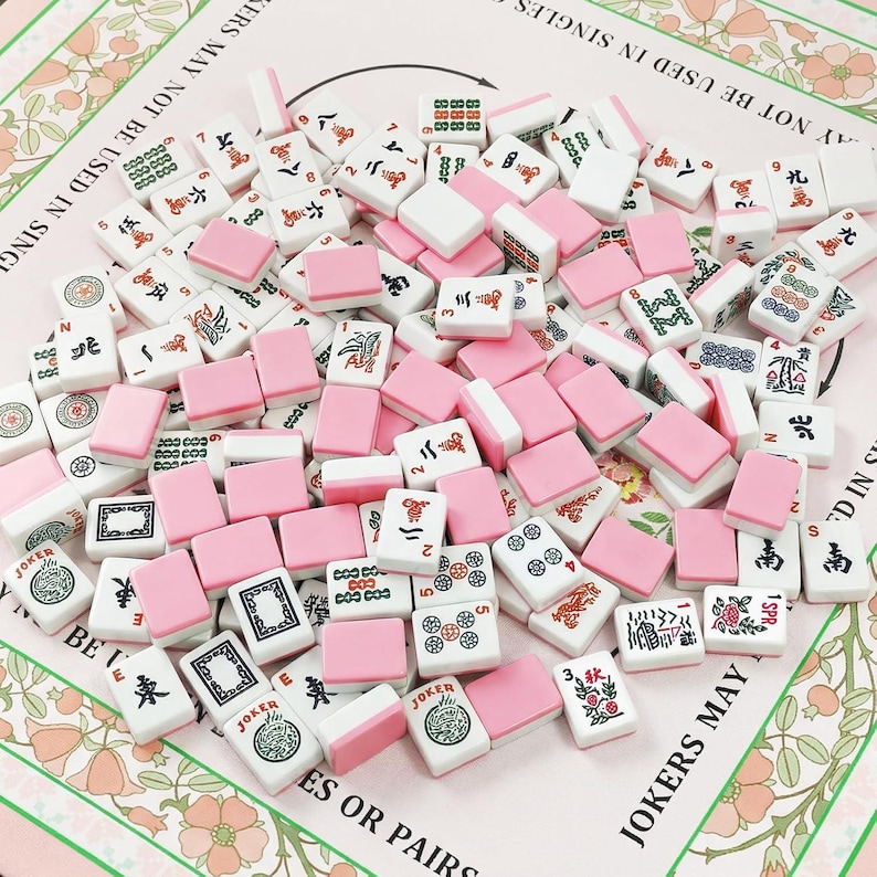 Pu&ograve; includere: Una collezione di tessere rettangolari da Mahjong con facce bianche che presentano vari simboli, caratteri e numeri tradizionali in inchiostro nero, rosso e verde, abbinate a dorsi rosa a tinta unita. Le tessere sono sparse su una superficie floreale.
